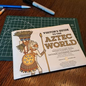 May include: A vintage "Visitor's Guide to the Aztec World" magazine cover featuring an illustration of an Aztec warrior. The magazine is on a green cutting mat, with a blue pencil, orange pencil, and black marker nearby.
