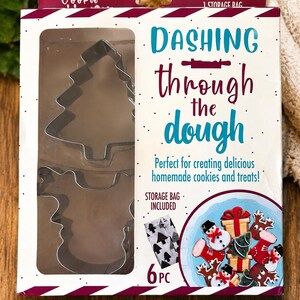 May include: A set of 5 metal cookie cutters and 1 storage bag in a white box with red striped edges. The box features the text "DASHING through the dough" and an illustration of decorated holiday cookies including snowmen, gifts, and reindeer.