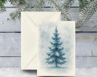 May include: A watercolor Christmas card featuring a blue evergreen tree with falling snow. The card is paired with a cream-colored envelope. The background is a light blue and white wooden surface with frosted pine branches.