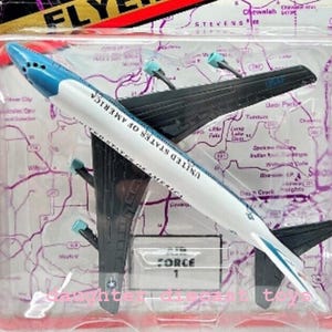 May include: A Road Champs Flyers toy aeroplane in a clear plastic package. The toy plane is white and blue with "UNITED STATES OF AMERICA" printed on the side. The packaging features a map design and the text "AIR FORCE 1".