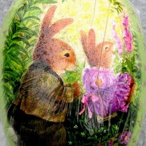 Könnte beinhalten: Ein bemaltes Osterei mit einer Szene von zwei Hasen in einem Garten. Ein Hase schaukelt den anderen. Das Ei ist grün mit einem Blumendesign und der Signatur des Künstlers.