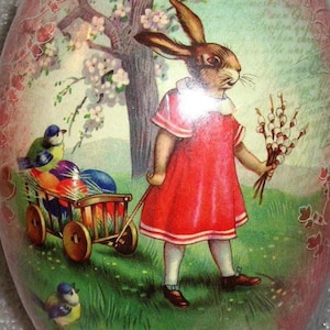 May include: An Easter egg with a painted scene of a rabbit-child in a red dress pulling a wagon of colorful eggs. The egg has a pink and green floral border and a tree with blossoms.