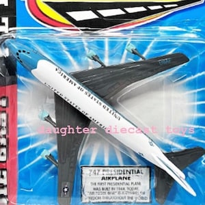 May include: A Road Champs diecast model of a 747 Presidential Aikplane. The toy airplane is white and blue with black wings and the text "UNITED STATES OF AMERICA". The packaging includes the text "AGES 4 & UP".