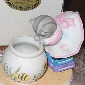 May include: A ceramic figurine of a gray tabby kitten peering into a white fishbowl. The kitten wears a pink and white dress with a large bow. The fishbowl has a yellow and black fish design.