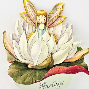 May include: A whimsical greeting card featuring a fairy figurine nestled in a white lotus flower. The fairy has golden wings and hair, wearing a blue dress. The card includes the word "Greetings" in script.