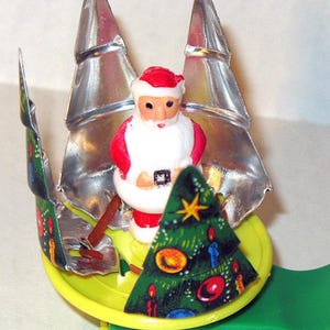 May include: A vintage Christmas ornament featuring Santa Claus in a sleigh. The sleigh is green and yellow, with silver tree-shaped accents. Santa is wearing a red and white suit, and a decorated Christmas tree is in front of him.