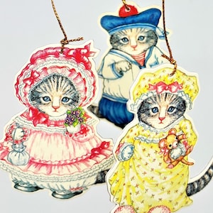 May include: Three vintage-style cat ornaments. One cat wears a pink dress and bonnet, another a sailor outfit, and the third a yellow nightgown and bonnet. Each cat has blue eyes and is holding an item.