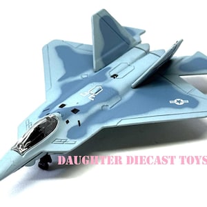May include: Diecast model of a light blue and gray fighter jet with a camouflage pattern. The toy plane has a detailed cockpit and wings, and the text "DAUGHTER DIECAST TOYS" is printed on the side. The model is set against a white background.