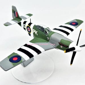 Puede incluir: Maqueta detallada de avión en tonos verdes, grises y blancos, con alas a rayas blancas y negras. El modelo presenta una hélice, una cabina transparente y marcas, incluyendo "PK" y una escarapela. Está montado sobre un soporte transparente.