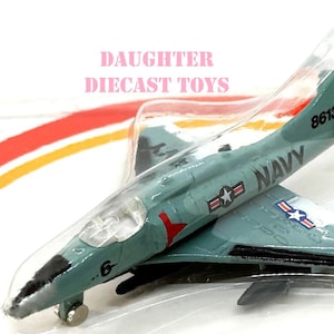 Puede incluir: Un modelo de avión de fundición a presión en un embalaje de plástico transparente. El avión de color verde azulado tiene la palabra "NAVY" impresa en el lateral, junto con el número "8613" y el texto "DAUGHTER DIECAST TOYS". Las alas presentan emblemas de estrellas.