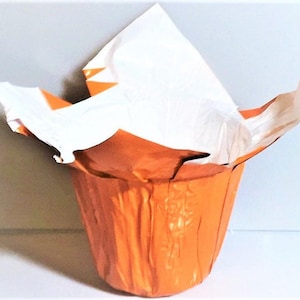 Könnte beinhalten: Ein orangefarbener, tulpenförmiger Behälter mit weißem Papier im Inneren. Der Behälter hat ein strukturiertes, metallisches Aussehen. Das weiße Papier hat orangefarbene Akzente und ist über die Oberseite gefaltet. Der Behälter steht auf einer weißen Oberfläche.