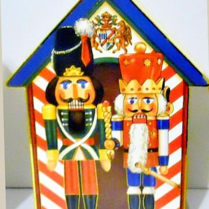 Könnte beinhalten: Dekorative Weihnachtsdekoration aus Holz mit zwei Nussknacker-Soldaten in einem hausförmigen Rahmen. Die Nussknacker tragen bunte Uniformen mit goldenen Akzenten. Das Haus hat eine rot-weiß gestreifte Fassade und ein blaues Dach.