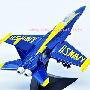 May include: A diecast model of a U.S. Navy jet aircraft. The model is primarily blue with yellow accents and the words "U.S. NAVY" in yellow. The model is mounted on a black stand.