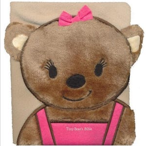 May include: A plush, brown teddy bear with a pink bow and a pink dress. The bear has black eyes and a black nose. The text "Tiny Bear's Bible" is printed on the dress. The bear is set against a beige background.