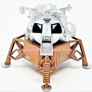 Op de afbeelding: Een gegoten model van de Apollo Lunar Module, met een zilveren bovenkant en een goudkleurige basis. De basis heeft de tekst "UNITED STATES" en een kleine Amerikaanse vlag. Het model heeft gedetailleerde kenmerken, waaronder landingsgestel en een ladder.