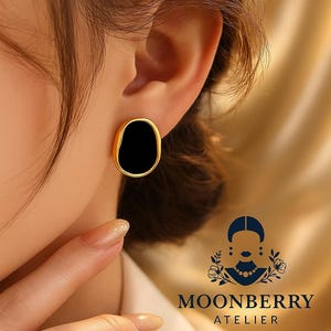 Puede incluir: Pendientes de botón dorados con un centro de esmalte negro. Los pendientes tienen una forma ovalada orgánica. La imagen también muestra el logotipo de Moonberry Atelier.