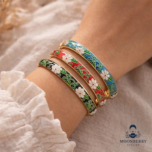 Può includere: Quattro braccialetti smaltati colorati con disegni floreali. I braccialetti sono dorati e presentano fiori bianchi con sfondi rossi, verdi, neri e blu. I braccialetti sono impilati su un polso.