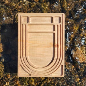 May include: A rectangular wooden serving board with a light, natural finish. The board features carved grooves and a curved design, likely for food presentation. The board is placed on a surface of water with rocks.