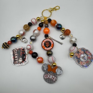 May include: A collection of San Francisco Giants-themed keychains. Each keychain features a gold clasp, colorful beads, and various charms, including baseballs, team logos, and a Mickey Mouse-shaped charm with the team's name.