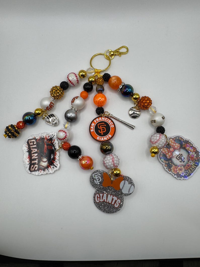May include: A collection of San Francisco Giants-themed keychains. Each keychain features a gold clasp, colorful beads, and various charms, including baseballs, team logos, and a Mickey Mouse-shaped charm with the team's name.
