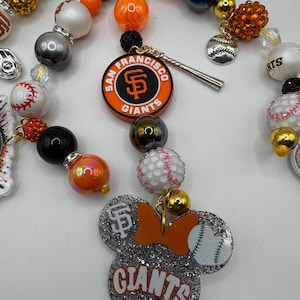 May include: A beaded necklace featuring San Francisco Giants baseball team charms. The necklace includes orange, black, and white beads, baseball-themed charms, and a Mickey Mouse-shaped charm with the Giants logo and baseball.