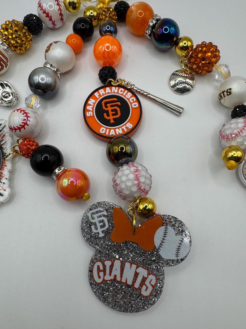 May include: A beaded necklace featuring San Francisco Giants baseball team charms. The necklace includes orange, black, and white beads, baseball-themed charms, and a Mickey Mouse-shaped charm with the Giants logo and baseball.
