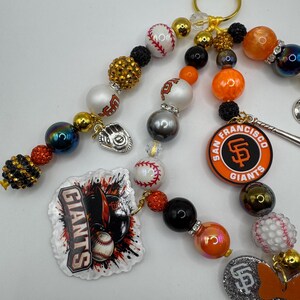 May include: Baseball-themed beaded keychain featuring San Francisco Giants logos, baseballs, and a baseball glove charm. The keychain includes gold, black, orange, and white beads, with a gold clasp. The Giants logo is displayed on a round charm.