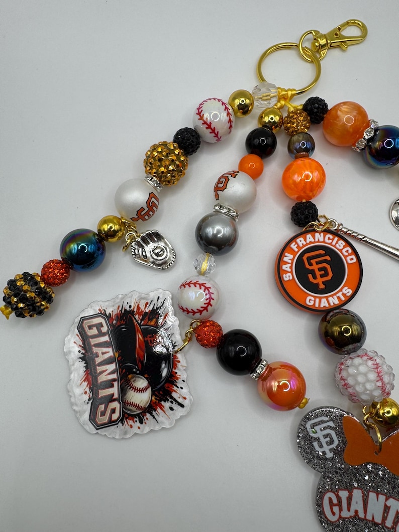 May include: Baseball-themed beaded keychain featuring San Francisco Giants logos, baseballs, and a baseball glove charm. The keychain includes gold, black, orange, and white beads, with a gold clasp. The Giants logo is displayed on a round charm.