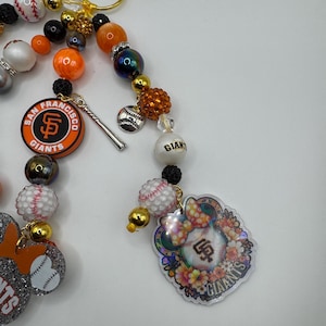 May include: A beaded keychain featuring San Francisco Giants baseball team charms. The keychain includes orange, black, white, and gold beads, baseball-themed charms, and a team logo. A gold-colored clasp is attached.