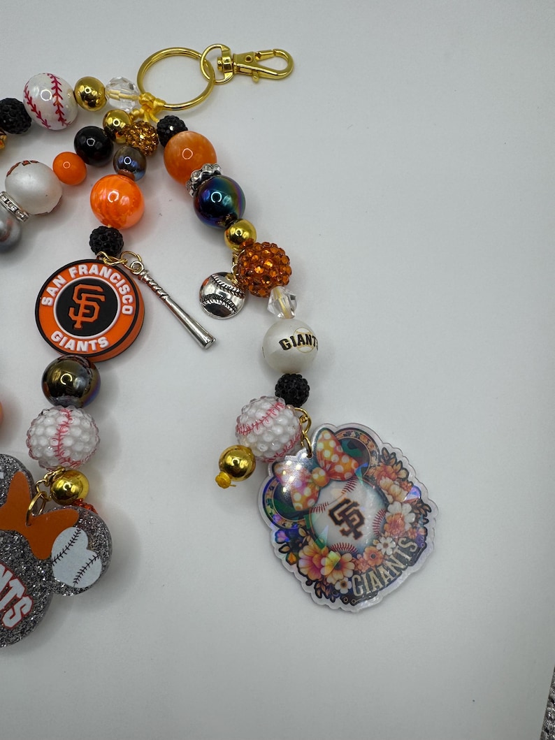 May include: A beaded keychain featuring San Francisco Giants baseball team charms. The keychain includes orange, black, white, and gold beads, baseball-themed charms, and a team logo. A gold-colored clasp is attached.
