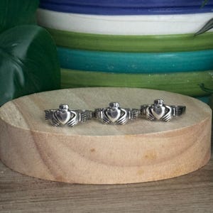 May include: Three silver-colored Claddagh rings are displayed on a round wooden surface. Each ring features a heart held by two hands, topped with a crown. The background includes a colorful ceramic pot and green foliage.