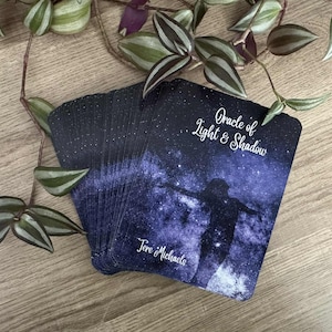 May include: A deck of oracle cards with a celestial design. The cards feature a dark blue background with a silhouette of a person with outstretched arms. The text "Oracle of Light & Shadow" and "Tere Michaels" are visible.
