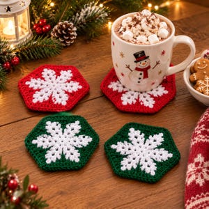 Könnte beinhalten: Ein Set aus vier hexagonalen Häkeluntersetzern mit weißen Schneeflockenmustern auf rotem und grünem Hintergrund. Auf einem Untersetzer steht eine weiße Keramiktasse, gefüllt mit heißer Schokolade, Marshmallows und einem Schneemann.