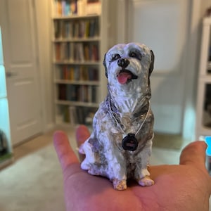 May include: A small, hand-painted dog figurine with a white and grey coat, black ears, and a pink tongue. The dog wears a black tag around its neck. The figurine is held in a person's hand.