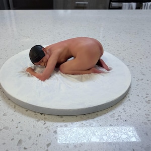 Könnte beinhalten: Eine nackte Skulptur einer Person, die auf einer weißen, kreisförmigen Plattform kniet. Die Figur befindet sich in einer Fötusstellung mit einer schwarzen Kappe. Die Plattform hat einen grauen Rand und steht auf einer gesprenkelten weißen Oberfläche.
