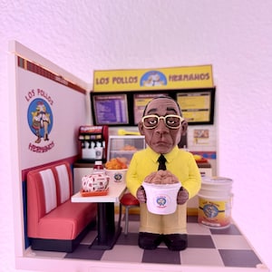 May include: A detailed miniature diorama depicting a Los Pollos Hermanos restaurant. A figure in a yellow shirt and glasses holds a bucket of food. The scene includes a booth, counter, and signage with the restaurant's logo and name.