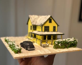 May include: A miniature model of a yellow house with a black car parked in front. The house has a porch and a snow-covered roof. The model is set on a wooden base with green bushes covered in snow.