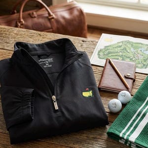 Op de afbeelding: Zwarte golfjas met een gouden rits en het Masters-logo. De afbeelding toont ook golfballen, een groen-witte gestreepte handdoek, een leren notitieboek en een golfbaan kaart op een houten tafel.