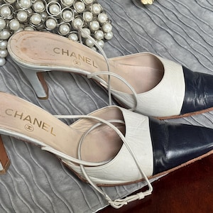 May include: A pair of Chanel mules featuring a two-tone design with white leather bodies and dark navy blue pointed toes. These slip-on shoes include thin, delicate ankle straps and a mid-height heel, with the Chanel logo printed on the inner sole.