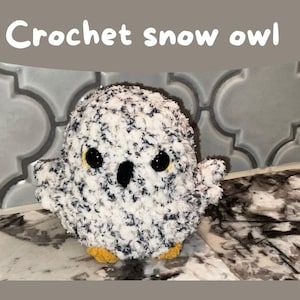 Może przedstawiać: Szydełkowa sowa śnieżna z puszystym białym korpusem nakrapianym ciemnoniebieskim. Ma duże, okrągłe czarne oczy z żółtymi akcentami i małe żółte stopy. Tekst "Crochet snow owl" znajduje się nad sową.