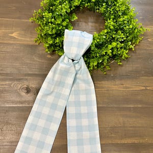 May include: A green, leafy wreath with a light blue and white gingham ribbon. The wreath is circular and appears to be made of artificial greenery. The ribbon is tied in a bow and hangs down from the wreath.