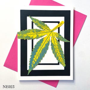 Puede incluir: Tarjeta de felicitación con una gran hoja de cannabis amarilla y azul estilizada. La hoja está centrada en una tarjeta blanca con un marco blanco y negro, sobre un sobre rosa. El texto "NE023" está en la esquina inferior izquierda.
