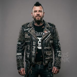 Puede incluir: Chaqueta de cuero negro con tachuelas plateadas y parches con nombres de bandas e imágenes de punk rock. La chaqueta tiene un estilo motero clásico con cuello de solapa y cinturón. La persona lleva una camiseta negra con un gráfico de calavera.