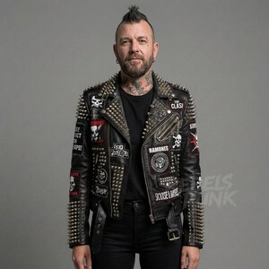 Op de afbeelding: Zwarte leren jas versierd met zilveren spikes en patches met bandlogo's en schedelmotieven. De jas heeft een ritssluiting, een riem en een ketting. Patches met "The Clash" en "Ramones".