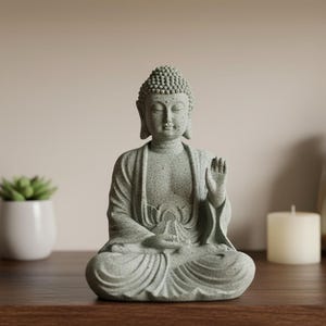 Op de afbeelding: Een zittend Boeddha-beeld in een meditatieve houding, gemaakt van gespikkelde grijze steen. De figuur heeft een serene uitdrukking, met één hand geheven in een gebaar van zegen. Het beeld is een decoratief item.