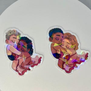 Könnte beinhalten: Zwei holografische Aufkleber mit Illustrationen von sich umarmenden Figuren. Die Figuren haben unterschiedliche Hauttöne und sind teilweise bekleidet. Die Aufkleber haben einen schimmernden, irisierenden Effekt und sind vor weißem Hintergrund.