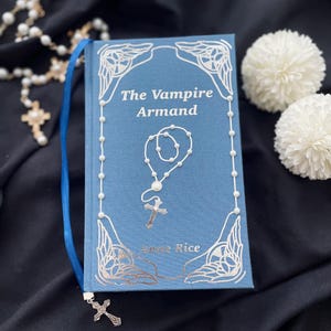 May include: A blue book titled "The Vampire Armand" by Anne Rice, with a white rosary and a blue ribbon bookmark. The book has decorative silver accents and is set against a dark background with white floral accents.