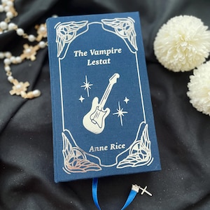 May include: A hardcover book titled "The Vampire Lestat" by Anne Rice. The book has a dark blue cover with silver foil accents, including a guitar illustration and decorative borders. A blue ribbon bookmark and a silver cross charm are attached.