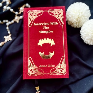 May include: A red hardcover book titled "Interview With The Vampire" by Anne Rice. The cover features gold foil accents and a stylized vampire teeth graphic. A black bookmark with a gold cross hangs from the book.