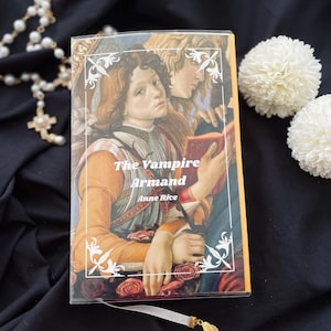 May include: A hardcover book titled "The Vampire Armand" by Anne Rice. The cover features a Renaissance-style painting of two figures. A white ribbon bookmark with a gold leaf charm is visible. The book rests on a black surface with white flowers and a beaded necklace.
